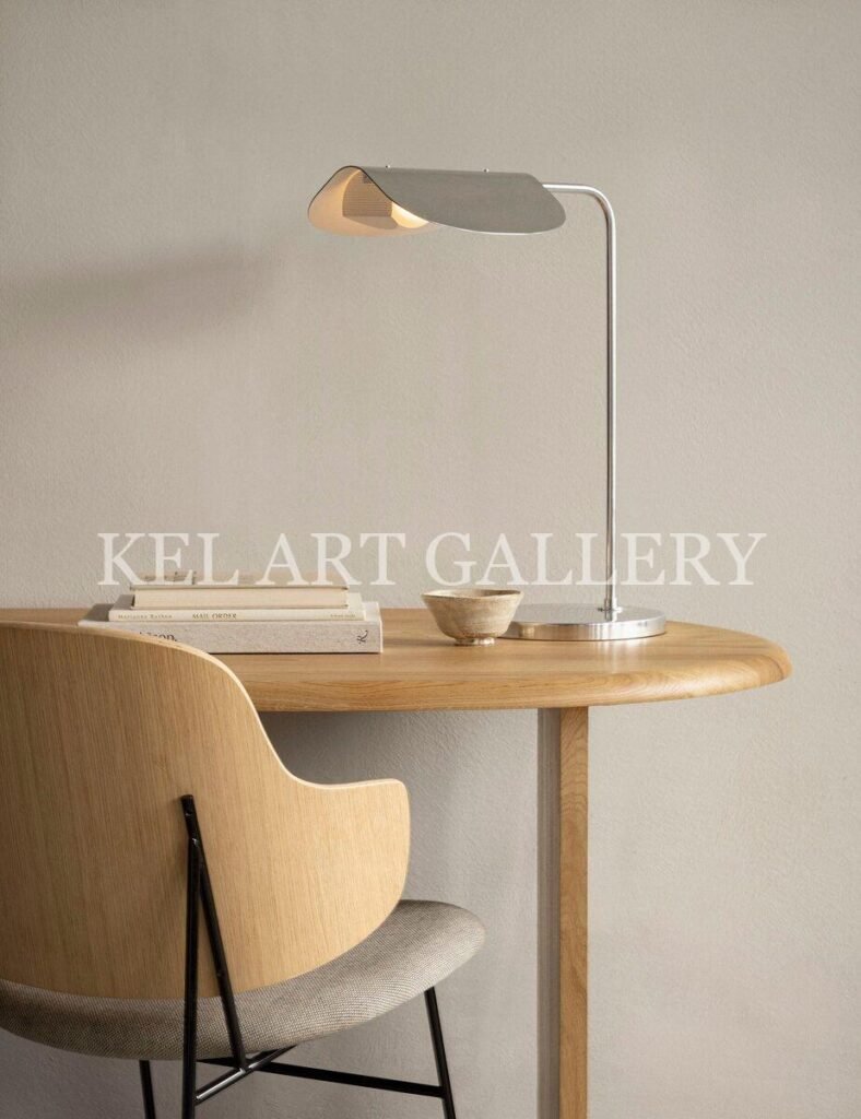 Minimalist Scandinavian-style wooden desk setup with a modern metal desk lamp, a curved wooden chair with fabric seat, a small ceramic bowl, and stacked neutral-toned books against a light beige wall.